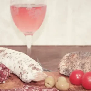 Planche en bois avec saucisson créole, olives, pain et tomates, accompagné d’un verre de vin rosé